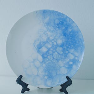 May include: A round white ceramic plate featuring a blue bubble-like pattern concentrated on the right side. The design consists of various circular shapes in shades of blue that fade into the plain white surface of the plate.