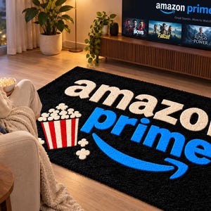 Könnte beinhalten: Ein rechteckiger schwarzer Teppich mit dem weißen Schriftzug "amazon" über dem blauen Schriftzug "prime" und einem geschwungenen Pfeil darunter. Links neben dem Text befindet sich eine Grafik eines rot-weiß gestreiften Popcorneimers.