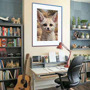 Op de afbeelding: Een ingelijste wandkunstprint met een portret van een woestijnvos met grote oren en lichtbruine vacht, tegen een woestijnachtergrond. Het kunstwerk hangt in een thuiskantoor met een bureau, een gitaar en boekenplanken vol met diverse voorwerpen.