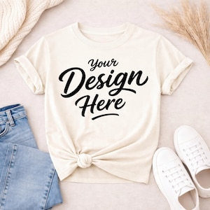 Könnte beinhalten: Cremefarbenes T-Shirt mit umgekrempelten Ärmeln und geknotetem Saum. Der Schriftzug "Your Design Here" ist in schwarzer Schrift aufgedruckt. Blaue Jeans und weiße Turnschuhe deuten auf einen lässigen, alltagstauglichen Stil hin.