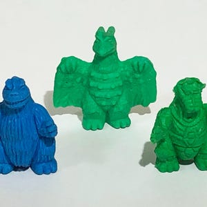 Può includere: Tre piccole figurine di mostri colorati. Un mostro blu con corpo rotondo e un mostro verde dall'aspetto di rettile sono ai lati di un mostro verde alato. I giocattoli sono fatti di un materiale solido.