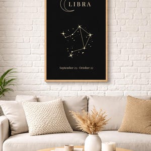 Peut inclure: Une affiche encadrée du signe du zodiaque Balance. Le fond noir présente un motif de constellation doré, le mot "LIBRA" et les dates "23 septembre - 22 octobre". L'affiche est affichée au-dessus d'un canapé beige avec des coussins décoratifs et une table basse en bois.