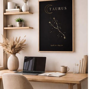 Peut inclure: Affiche noire encadrée avec texte doré et un motif de constellation, intitulée "TAURUS" et "April 20 - May 20." L'affiche est au-dessus d'un bureau en bois avec un ordinateur portable, un livre ouvert et une tasse beige.