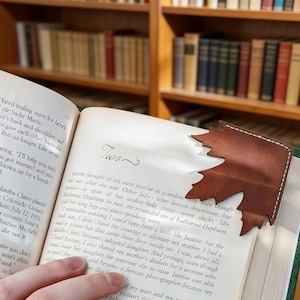 May include: A brown leather leaf-shaped bookmark with white stitching, placed on the corner of a book. The bookmark's design resembles a maple leaf. The book is open to a page with printed text.