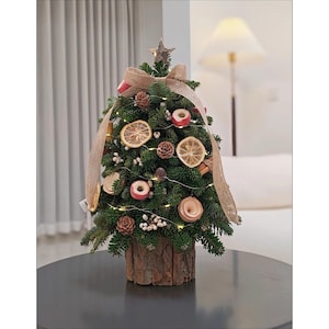 Op de afbeelding: Een kleine, versierde kerstboom met een rustieke houten basis. De boom is versierd met gedroogde sinaasappelschijfjes, dennenappels en kleine rode ornamenten. Een jute lint en een ster bekronen de boom, met kleine lichtjes.