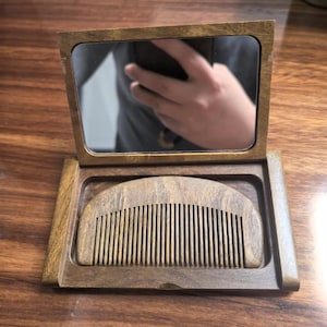 May include: A wooden travel comb and mirror set. The comb sits in a tray, and the mirror is hinged to stand upright. The wood has a warm, natural tone. The mirror reflects a person holding a phone.