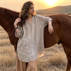 May include: A beige, long-sleeved, loose-fitting tunic top with a V-neckline and a curved hemline. The garment features a simple, unpatterned design with a relaxed silhouette, mid-length sleeves, and side slits, crafted from a textured fabric.