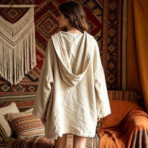 May include: A beige hooded jacket with long sleeves. The jacket is made of a lightweight fabric and has a relaxed fit. The model is wearing the jacket in a room with patterned textiles and a sofa.