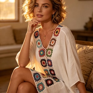 May include: A white, V-neck dress with colorful crochet detailing. The dress features a floral pattern in shades of blue, red, yellow, and green. The model is sitting on a couch, wearing a necklace and bracelets.