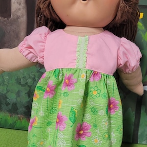 Pode incluir: Uma boneca vintage com cabelo de fio castanho e um rosto redondo. A boneca usa um top rosa com acabamento de babados e um vestido verde com estampa floral. O vestido apresenta flores rosa e amarelas. Os braços e pernas da boneca são de material bege.