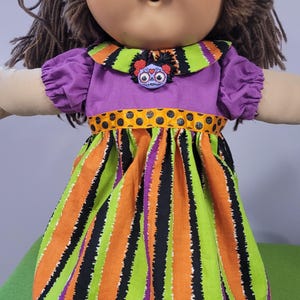 Puede incluir: Una muñeca con cabello de lana marrón y cara redonda que viste un vestido morado con un cuello que presenta un broche de calavera. El vestido tiene una cintura con lunares negros y naranjas y una falda con rayas verticales naranjas, verdes y negras.