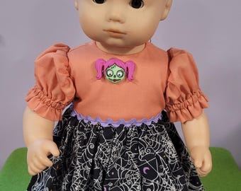 May include: A doll with dark hair wearing a dress that has a solid peach-colored bodice with a small green zombie-like character embellishment and a black skirt featuring a white spiderweb pattern with small colorful stars and crescent moons.