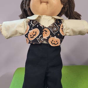 May include: A doll with brown yarn hair and blue eyes wearing a cream-colored long-sleeve shirt, a black vest featuring an orange pumpkin and owl print, and solid black pants. The doll has a rounded face with freckles and visible toes.