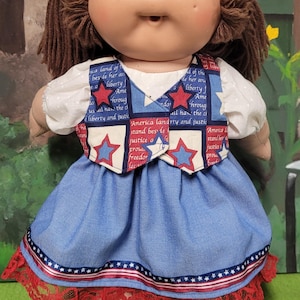 Op de afbeelding: Een vintage pop met bruin haar en een lichte huidskleur, gekleed in een patriottische outfit. De blauwe jurk van de pop heeft een rood-witte bies en een vest met sterren en tekst. De outfit doet denken aan de Amerikaanse vlag.