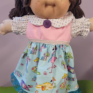 May include: A doll with brown yarn hair and blue eyes wearing a dress with a pink bodice, a white collar with colorful dots, and a light blue skirt featuring a print of pink flamingos, palm trees, and beach items, finished with teal lace.