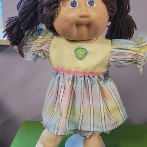 May include: A doll with brown yarn hair styled in pigtails, blue eyes, and a round face with freckles. The doll wears a yellow bodice featuring a green heart appliqué, a pink rickrack trim, and a pastel plaid skirt with white lace trim.