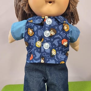 May include: A doll with brown yarn hair and a tan complexion wearing a blue short-sleeved shirt under a button-down vest featuring a blue pattern of cartoon characters and magical symbols, paired with dark blue denim trousers.