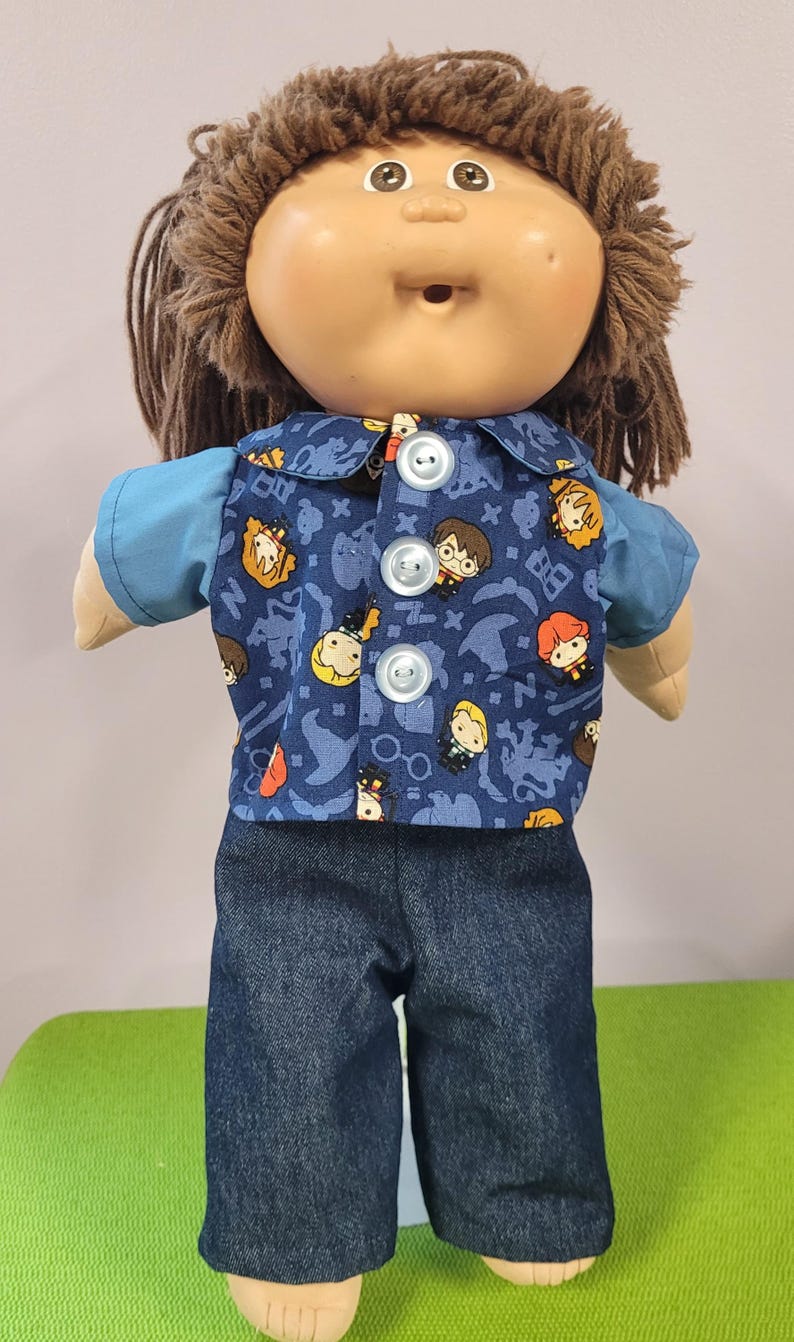 May include: A doll with brown yarn hair and a tan complexion wearing a blue short-sleeved shirt under a button-down vest featuring a blue pattern of cartoon characters and magical symbols, paired with dark blue denim trousers.