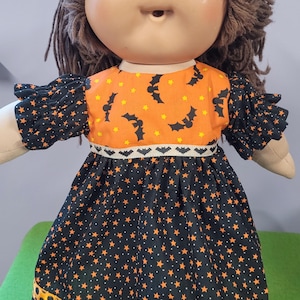 May include: A doll with brown yarn hair and a round face wearing a dress with an orange bodice featuring black bats and yellow stars. The black skirt has orange stars and a hem with orange and black polka dots, accented by a bat-patterned ribbon.