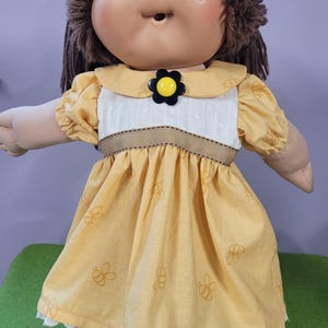 May include: A doll with brown yarn hair and a round face wearing a yellow dress with a bee print pattern. The dress features a white bodice, a brown ribbon waistband, and a black and yellow flower-shaped button at the center of the collar.