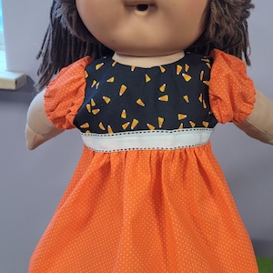 May include: A doll with brown yarn hair and a round face wearing a dress with a black bodice featuring a candy corn pattern, orange puff sleeves, and an orange skirt with white polka dots, a white ribbon waistband, and a black lace hem.