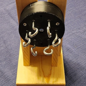 May include: A wooden stand with a black, circular top featuring six metal hooks. The stand is made of light-coloured wood and sits on a rectangular base. The hooks are evenly spaced around the circular top.