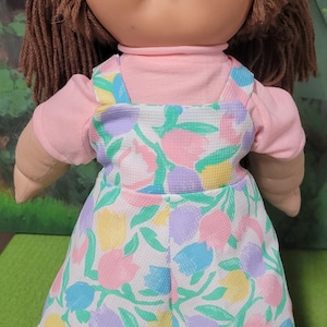 May include: A vintage Cabbage Patch Kid doll with brown yarn hair and a round face. The doll wears a pink long-sleeved shirt and a white romper with a floral pattern in pastel colors. The romper has a lace trim.