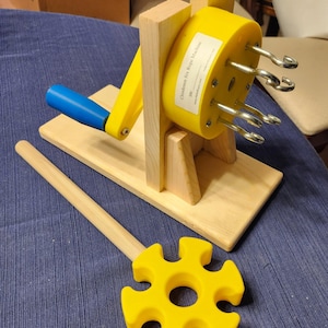 Może przedstawiać: Żółto-niebieska maszyna do robienia lin z drewnianą podstawą i uchwytem. Maszyna posiada metalowe haczyki i drewniane narzędzie z żółtym, kwiatowym zakończeniem. Tekst na maszynie brzmi "Clendenon Six Rope Machine".