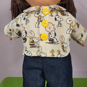 May include: A doll with brown yarn hair wearing a collared shirt featuring a yellow and white striped pattern with Snoopy and Charlie Brown graphics, fastened with three bright yellow buttons, paired with dark blue denim pants.