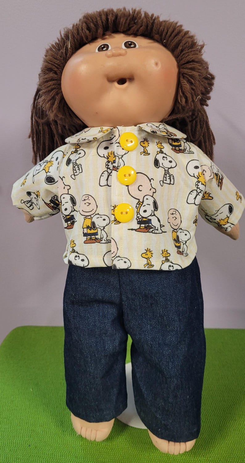 May include: A doll with brown yarn hair wearing a collared shirt featuring a yellow and white striped pattern with Snoopy and Charlie Brown graphics, fastened with three bright yellow buttons, paired with dark blue denim trousers.