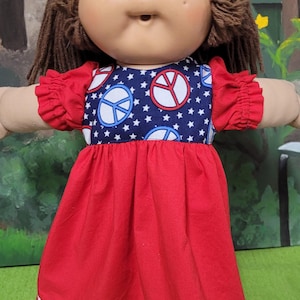 Könnte beinhalten: Eine Vintage-Puppe mit braunen Haaren und rundem Gesicht, die ein rotes Kleid trägt. Das Kleid hat ein marineblaues Oberteil mit weißen Sternen und Friedenszeichen. Das Kleid hat rote Ärmel und eine weiße und marineblaue Spitzenborte.