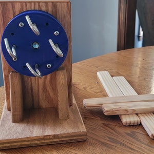 Puede incluir: Una rueda de hilar de madera azul con ganchos de metal sobre una base de madera. La rueda está montada en un soporte de madera con dos patas. También hay cuatro tablones de madera sobre la mesa.