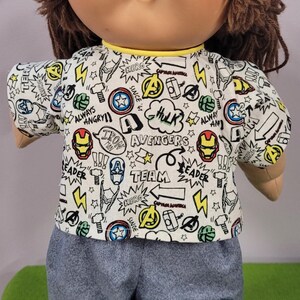 May include: A doll with brown yarn hair and a round face wearing a short-sleeved white shirt featuring a repeating Marvel Avengers comic book print with text including Iron Man, Hulk, and Team, paired with solid grey trousers with an elastic waist.