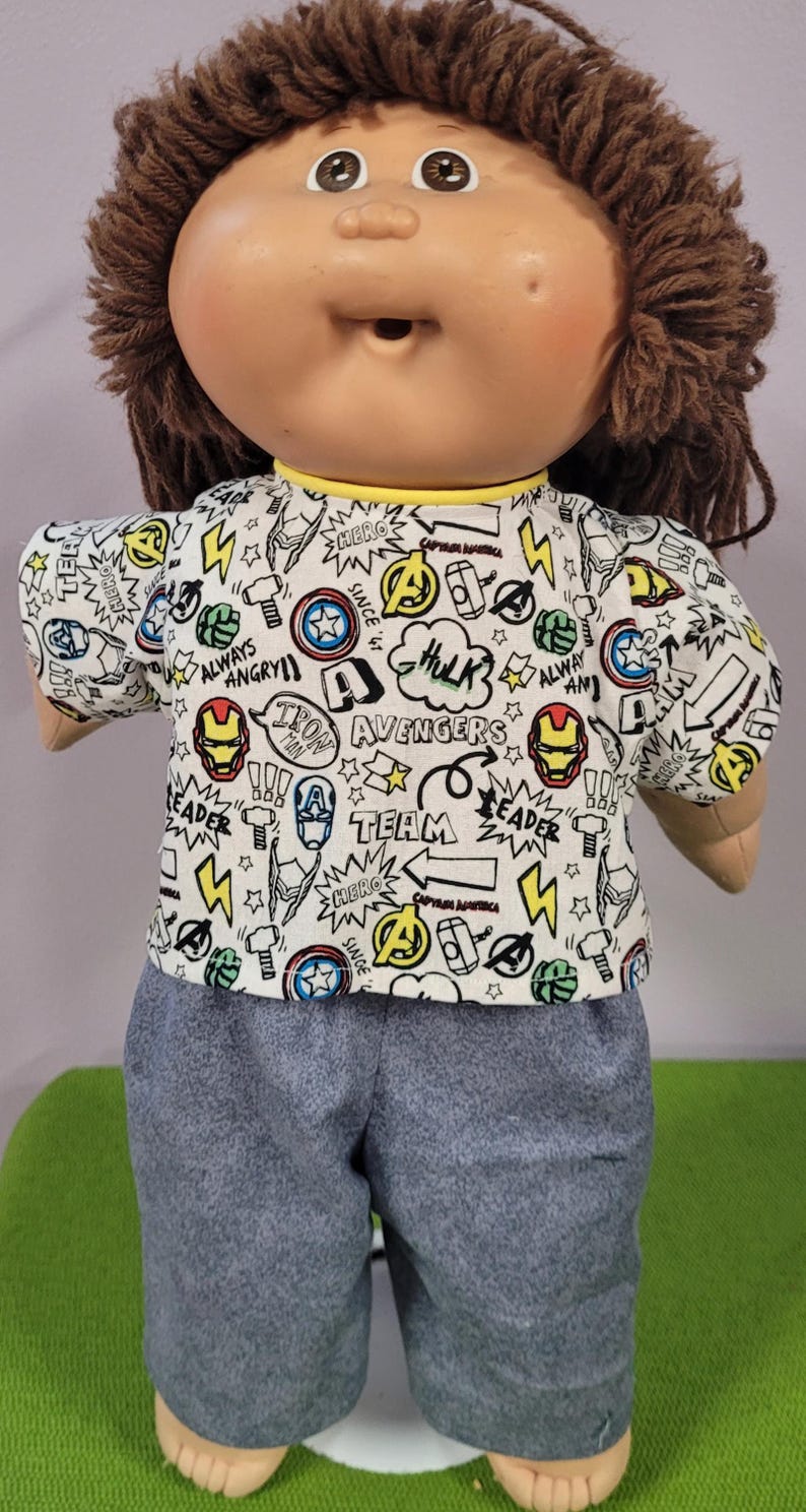 May include: A doll with brown yarn hair and a round face wearing a short-sleeved white shirt featuring a repeating Marvel Avengers comic book print with text including Iron Man, Hulk, and Team, paired with solid grey trousers with an elastic waist.