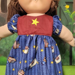 May include: A vintage doll with brown hair and a red and blue dress. The dress features a yellow star and a pattern of stars and patriotic imagery. The doll has a round face and is posed against a painted backdrop.