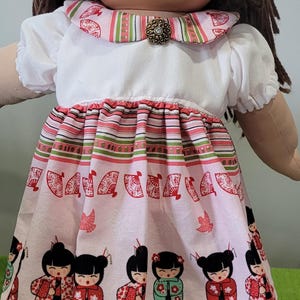 May include: A doll with brown yarn hair and a round face wearing a white short-sleeved dress. The dress features a pink skirt with a pattern of small figures in traditional clothing, red fans, and floral motifs, accented by a striped collar and a brooch.