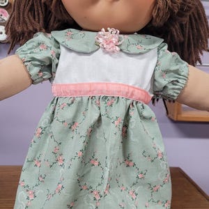 May include: A doll with brown yarn hair and a round face wearing a light green dress with a white bodice, a pink ribbon waistband, and a small pink flower on the collar. The dress features a pattern of tiny pink flowers and white ribbons.