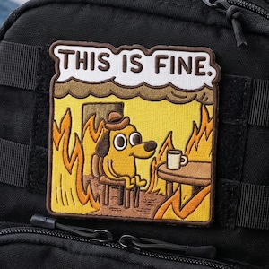 Könnte beinhalten: Schwarzer Rucksack mit einem Aufnäher mit der Aufschrift "THIS IS FINE." Der Aufnäher zeigt einen Cartoon-Hund in einem brennenden Raum, mit einer Kaffeetasse auf einem Tisch. Der Rucksack hat mehrere Riemen und Reißverschlüsse.