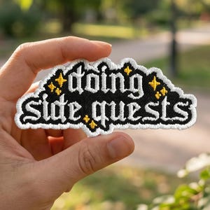 May include: Embroidered patch with the words "doing side quests" in white, stylized lettering on a black cloud-shaped background. The patch is outlined in white with gold star accents. The patch is held in a hand against a blurred green background.