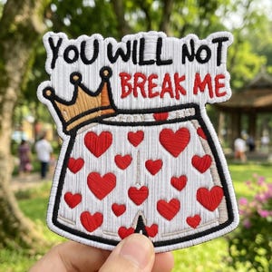 May include: Embroidered patch with the text "YOU WILL NOT BREAK ME" above a design of a crown and a pair of white shorts covered in red hearts. The patch has a black border.