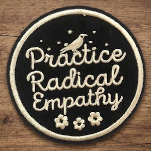 May include: A round, black embroidered patch with a white border. The patch features the words "Practice Radical Empathy" in a cursive font, along with a bird and three small flower designs. The patch is set against a wooden background.