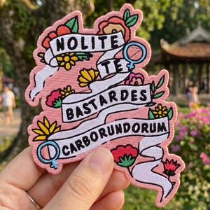 May include: A pink embroidered patch with the words "Nolite Te Bastardes Carborundorum" in black, white, and red. The patch features floral and gender symbol designs. The patch is held in a hand.