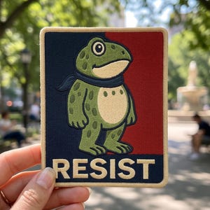 May include: Embroidered patch featuring a cartoon frog with a blue scarf, set against a red and navy blue background. The word "RESIST" is displayed in white block letters at the bottom. The patch is rectangular with a tan border.