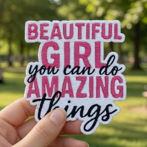 May include: A white patch with the words "BEAUTIFUL GIRL you can do AMAZING things" in pink and black lettering. The patch is held in front of a blurred green park background.