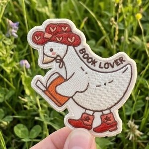 May include: An embroidered patch featuring a cartoon goose wearing a red hat and boots, holding a book. The text "BOOK LOVER" is printed on the patch. The patch is held in front of a background of green grass and wildflowers.