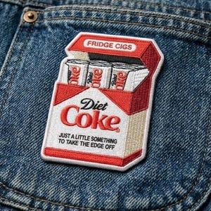May include: Embroidered patch featuring a Diet Coke cigarette pack design. The patch is red, white, and beige, with the words "Diet Coke" and "Fridge Cigs" printed on it. The patch is attached to a denim jacket.