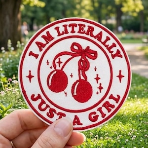 May include: A round white patch with red text and a cherry design. The text reads "I AM LITERALLY JUST A GIRL". The patch is held in a hand against a blurred park background.
