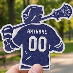 Op de afbeelding: Een marineblauw geborduurde patch met een lacrosse-speler met een lacrosse-stick. Het shirt van de speler toont de tekst "ANYNAME" boven het nummer "00" in het wit. De patch heeft een witte rand en wordt in een hand gehouden.