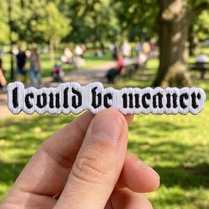 Może przedstawiać: Biała naszywka z czarnym napisem "I could be meaner". Naszywka ma czarną obwódkę i falisty brzeg. Tło to park z drzewami i ludźmi.