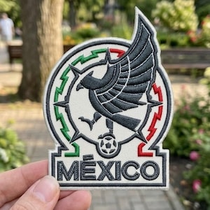 Puede incluir: Un parche bordado blanco que presenta un águila negra con las alas extendidas posada sobre un balón de fútbol. El diseño está enmarcado por un borde circular con detalles en verde y rojo y la palabra MEXICO en letras mayúsculas negras.